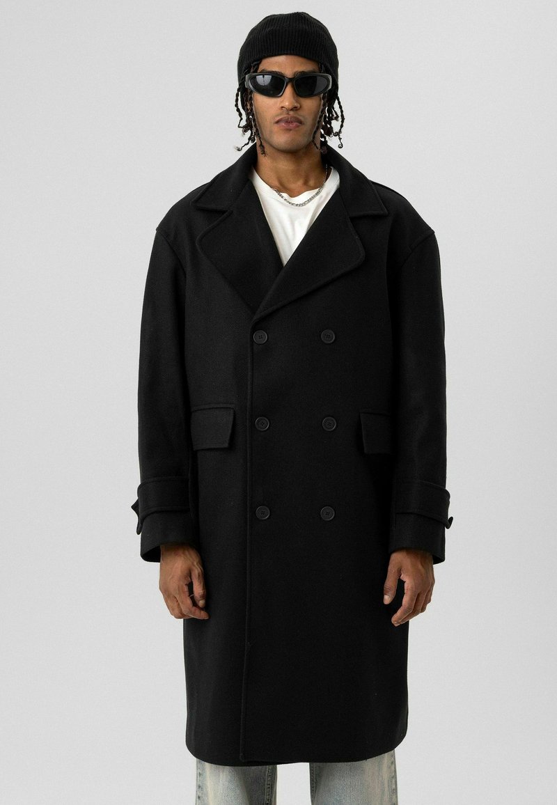 Persoon met een zwarte double-breasted jas, zwarte beanie, donkere zonnebril, wit overhemd, zilveren ketting, en lichtblauwe spijkerbroek, staand tegen een witte achtergrond.