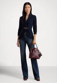 Modrý pletený sveter s opaskom v páse, doplnený tmavomodrými džínsami. Drží bordovú kabelku so zlatými kovaniami a strapcovými detailmi.