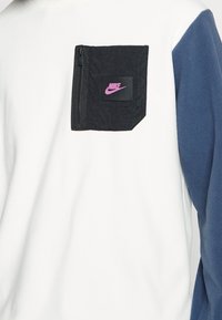Nike-hoodie med vit kropp, marinblå ärmar och en svart dragkedjeficka med en rosa logotyp. Tyget har en slät textur.