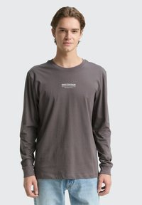 Pelēka kokvilnas krekla ar garām piedurknēm un apaļu izgriezumu, uz krūtīm tam ir balts logo teksta. Saskaņots ar gaiši zilu džinsu biksēm. Gluda tekstūra.