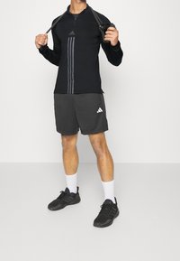 Schwarzes langärmliges Sportshirt mit grauen Streifen, kombiniert mit grauen Shorts mit weißem Adidas-Logo. Schwarze Sportschuhe und weiße Socken.