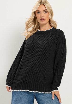 Donna bionda che indossa un maglione nero a maglia waffle con bordi bianchi smerlati e jeans blu su uno sfondo chiaro uniforme.