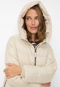 Beige dynejakke med stor, justerbar hætte, quiltet tekstur og lynlås foran. Ærmerne er brede med en blød elastisk manchet.