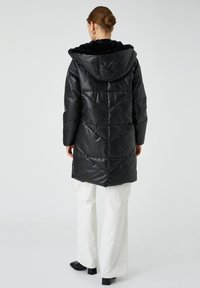 Vrouw met een haarbun, gekleed in een zwarte gewatteerde jas met een bontvoering, witte broek en zwarte hakken, staand tegen een effen achtergrond.