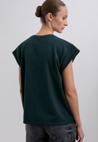 Dark green cotton t-shirt with rolled cap sleeves, a relaxed fit, and a rounded neckline, viewed from the back with minimal stitching details.