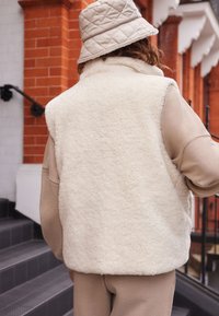 Gilet fluffy di colore crema realizzato in shearling, con un alto colletto e senza maniche, abbinato a una maglietta a maniche lunghe beige chiaro e un cappello a secchiello trapuntato.