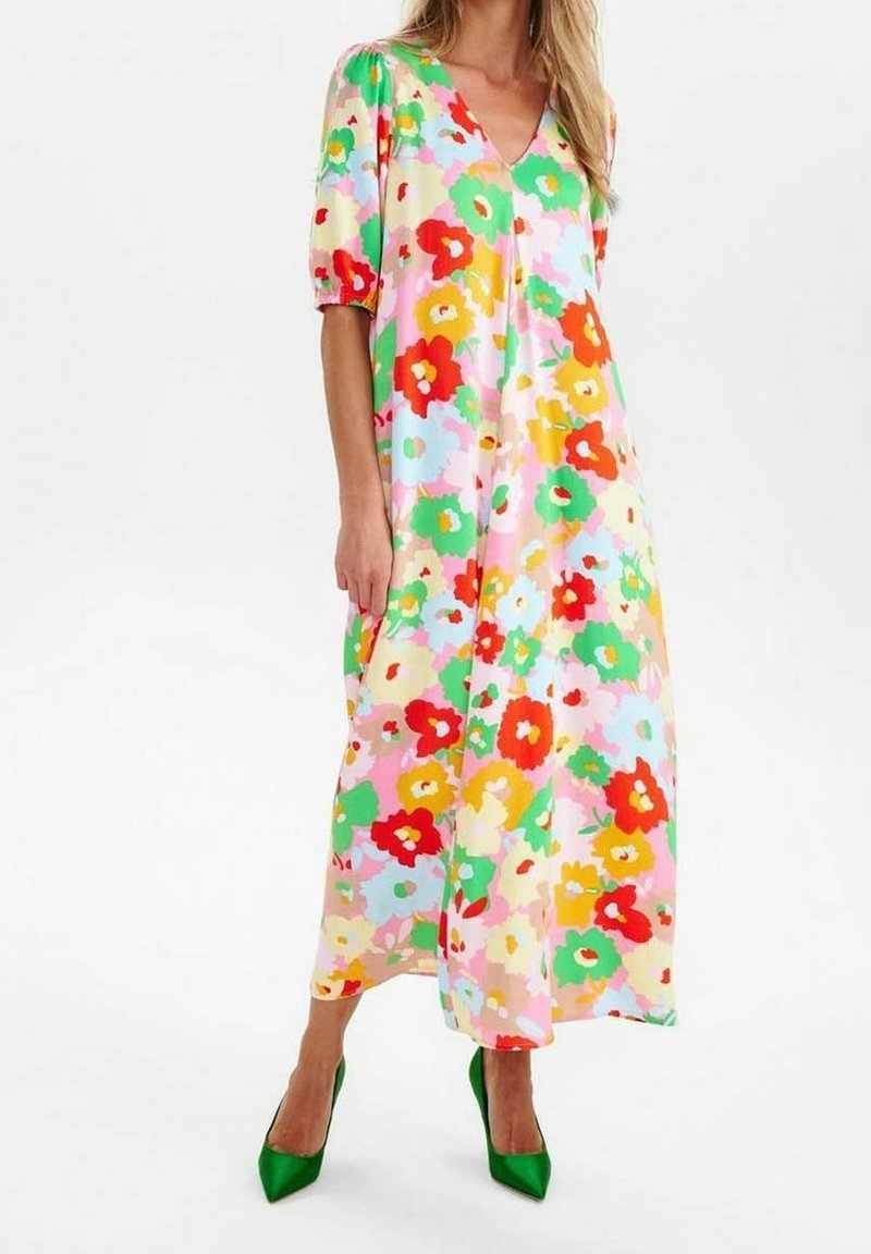 Femme portant une robe midi fleurie colorée avec des manches courtes bouffantes et des chaussures à talons verts, debout sur un fond blanc.