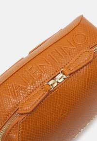 Orange texturerad läderväska med präglad logotyp, hårdvara i guldton och dragkedjestängning. Utrustad med en dekorativ dragkedjepull och slät yta.