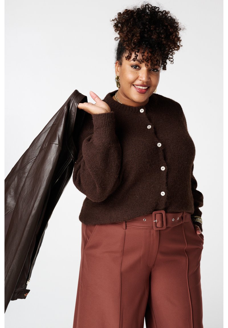 Cardigan en laine marron à encolure ronde, boutons sur le devant et coupe ample, associé à un pantalon taille haute couleur rouille et une veste en cuir marron.