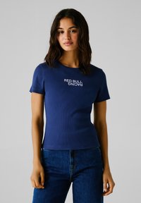 Camiseta ajustada de color azul marino con textura acanalada y mangas cortas, que presenta el texto en blanco "RED BULL RACING" en el frente. Combinada con jeans de mezclilla.