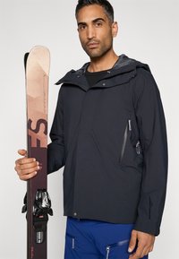 Casaco de esqui preto com bolso com fecho, forro cinzento e capuz, combinado com um par de esquis com um design gráfico e fixações.