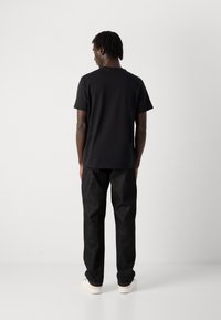 Man met korte dreadlocks staat met zijn rug naar de camera gekeerd, draagt een plain zwart t-shirt, donkere jeans en witte sneakers tegen een witte achtergrond.