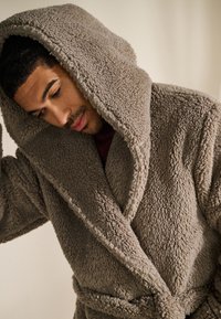 Peignoir à capuche beige douillet avec un tissu texturé et moelleux. Dispose d'un design croisé avec une ceinture à nouer et de grandes poches, offrant confort et chaleur.