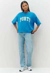 Moteris vilkinti mėlyną oversized "PORTO" džemperį, šviesiai mėlynas kelnes ir baltus sportinius batus, stovinti prieš paprastą šviesią foną.
