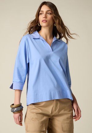 Kvinde iført lyseblå polo t-shirt med 3/4-ærmer og beige bukser, accessorized med store ringearringe og chunky armbånd.