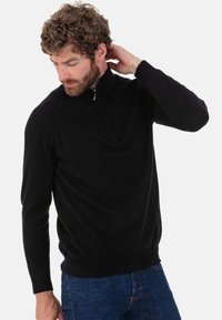 Schwarzer Pullover mit Reißverschluss am Kragen aus weichem Strickmaterial, mit langen Ärmeln und figurbetontem Schnitt, kombiniert mit dunkelblauen Jeans.