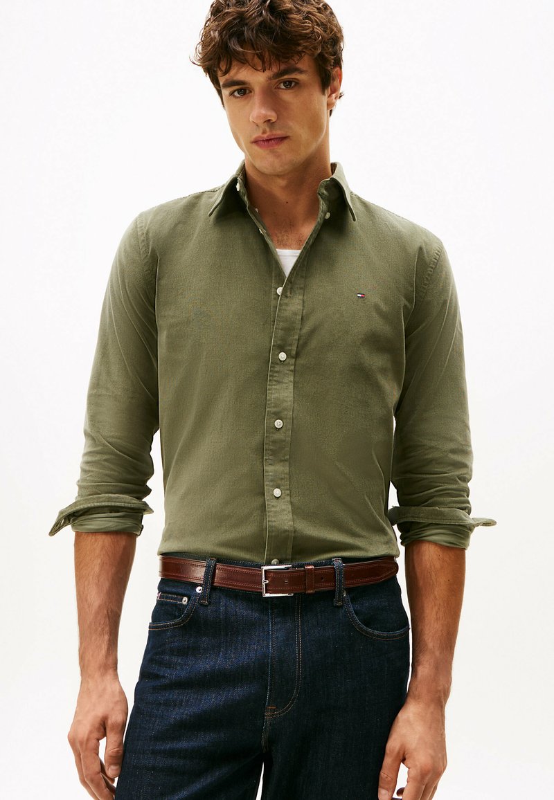 Camisa de manga larga color verde oliva con botones, que presenta un pequeño logo en el pecho, combinada con jeans azul oscuro y un cinturón marrón.