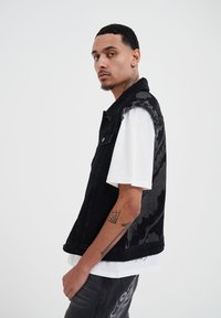 Gilet in denim nero con bordi sfrangiati, con un motivo floreale su una spalla. Indossato sopra una camicia bianca, con un tatuaggio visibile sul braccio.