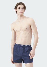 Tmavě modré boxerky s elastickým pasem a bílým logem. Materiál vypadá hladce, vpředu je detail s knoflíkem.