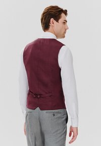 Bordowy lniany kamizelka o dopasowanym kroju. Tył z dwoma guzikami i subtelną teksturą, w zestawieniu z białą koszulą i szarymi spodniami w paski.