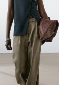 Tmavý top bez rukávů s asymetrickým střihem, světlé béžové široké kalhoty s proužky a hnědá texturovaná tkaná taška.