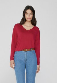 Rotes Langarm-V-Ausschnitt-Oberteil aus weichem Material, kombiniert mit hochgeschnittenen hellblauen Jeans und einem braunen Gürtel.