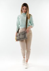 Donna con maglione azzurro chiaro e pantaloni beige che cammina, portando una borsa a tracolla grigia con portachiavi giallo e indossando scarpe da ginnastica bianche.