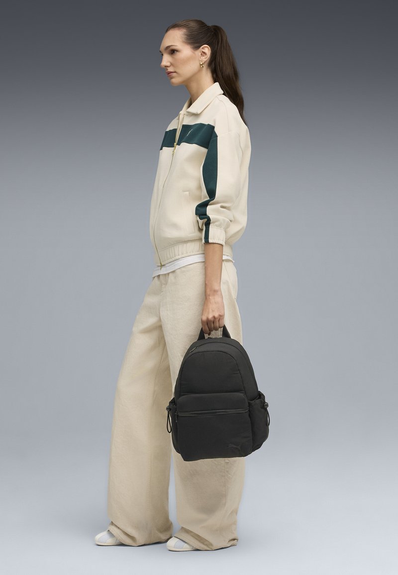 Un sac à dos noir avec une structure rembourrée et des poches latérales, associé à une veste crème avec des rayures vert foncé et un pantalon beige ample.