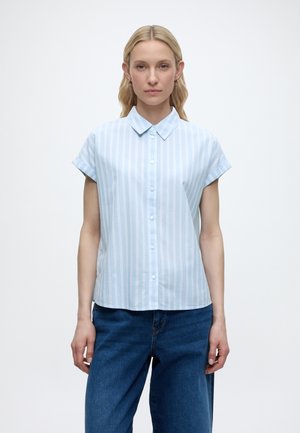 Femme portant une chemise à manches courtes bleu clair à rayures blanches verticales et un jean bleu, debout devant un fond blanc uni.