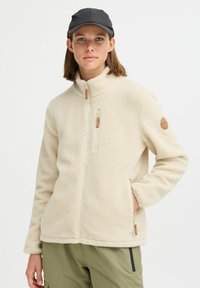 Veste en polaire beige clair avec un col haut, fermeture éclair sur le devant, poches latérales et un tissu texturé. Elle présente un écusson de logo beige sur la manche.