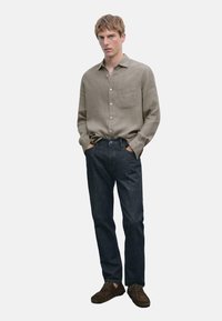 Light brown linen shirt with front pocket, paired with dark blue denim jeans and brown lace-up shoes. Model stands with hands in pockets.