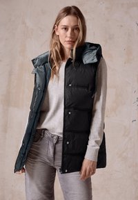 Schwarze gepolsterte Weste mit verstellbarem Kapuze, grauen Akzenten, Reißverschluss und Druckknopfverschluss sowieseitlichen Taschen, getragen über einem grauen Pullover.