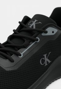 Artimatis juodas Calvin Klein sportinis batelis su tinklelio tekstūra, juodais raišteliais ir pilkais akytėmis, kuriame iš šono ir liežuvio matomas "cK" logotipas.
