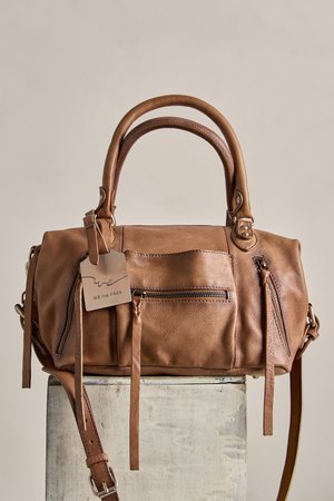 Καφέ δερμάτινη γυναικεία τσάντα με διπλές λαβές, εμπρόσθιες και πλαϊνές τσέπες με φερμουάρ, και μια ετικέτα που γράφει "We The Free," ακουμπισμένη σε ανοιχτό γκρι τετράγωνο.