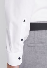 Vit skjort med svart ros- broderi på manschetten. Har mörka knappar och en slät textur. Tillsammans med ljusgrå byxor.