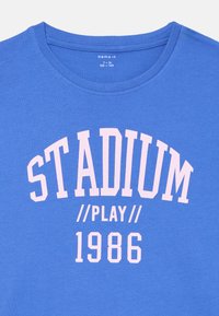 Modrá bavlněná tričko s kulatým výstřihem a růžovým potiskem textu: "STADIUM // PLAY // 1986" v obloukovém designu. Hladký povrch.