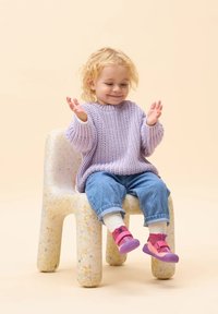 Jasnofioletowy sweter dzianinowy, niebieskie dżinsy oraz różowo-fioletowe buty. Dziecko siedzi na nakrapianym krześle na tle miękkiego beżowego koloru.