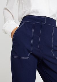 Bílá dlouhá košile s dlouhými rukávy zasunutá do tmavě modrých kalhot s vysokým pasem, které mají bílý kontrastní šev, a ruka v kapse.