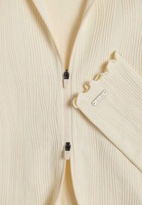 Giacca zip con una finitura cremosa e linee verticali testurizzate, guanti senza dita a coste coordinati. Presenta una zip nera e un dettaglio del logo argentato.
