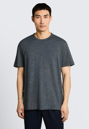 Młody mężczyzna z krótkimi czarnymi włosami, ubrany w jednolitą ciemnoszarą koszulkę z krótkim rękawem i okrągłym dekoltem na jasnoszarym tle.