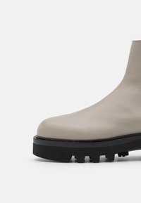 Botín de tobillo de cuero beige con un acabado suave, punta redondeada y suela externa de goma negra gruesa con un patrón texturizado de agarre.