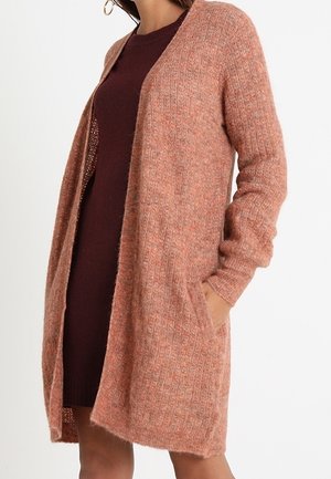 Nainen, joka pukeutuu pitkään, avoimeen vaaleanpunaiseen, teksturoituun neuletakkiin tumman viininpunaisen vartalonmyötäisen mekon päällä, toinen käsi neuletakin taskussa.