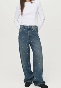 Wide-leg denim jeans with a dark wash, featuring a textured pattern of small circles and silver accents, styled with a white long-sleeve top.