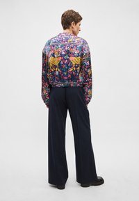 Veste bomber colorée présentant un motif floral et animalier aux teintes vives, associée à un pantalon sombre à fines rayures et des chaussures noires massives.