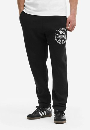 Mężczyzna w czarnych spodniach dresowych Lonsdale z białym logo na lewej udzie oraz czarnych trampkach Adidas z białymi paskami, stojący na tle białej ściany.