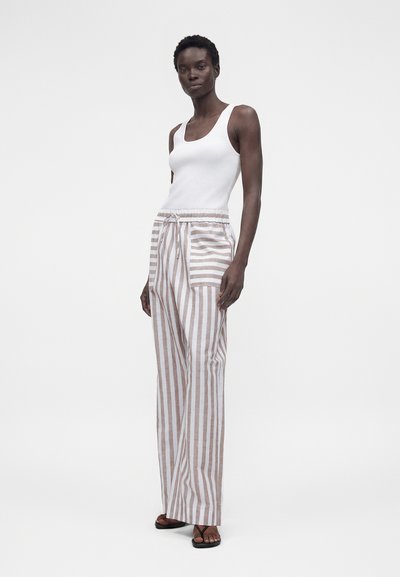 Γυναίκα στέκεται φορώντας λευκό τοπ χωρίς μανίκια και μπεζ-άσπρο ριγέ παντελόνι με ψηλή μέση και φαρδιά πόδια, με εμπρόσθιες τσέπες.
