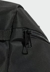 Čierny látkový roh batohu s vreckom na zips a textúrovaným povrchom, s hladkým tmavým jazdcom na zips a detailom popruhu.