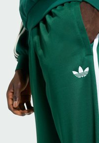 Zelené sportovní tepláky z hladkého materiálu, s bílým logem Adidas a bílými bočními pruhy. Obsahují elastický pás a detail kapsy.