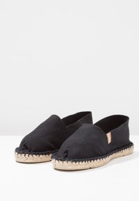 Black canvas espadrilles with a round toe, jute rope sole, and stitched detailing. No laces, featuring a small brown tag on the side.