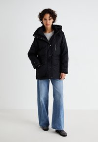 Svart pufferjacka med huva, tryckknappar, justerbar dragsko i midjan och sidofickor. Bärs över en grå topp och matchas med blå jeans.